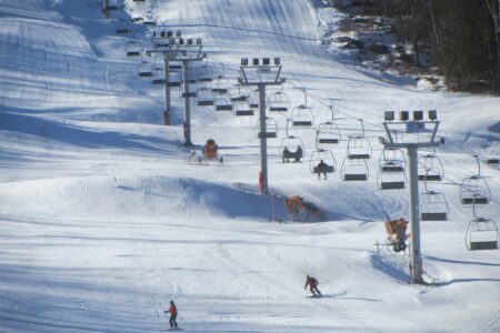 Skiers glide gracefully down the snowy slope of West Mountain, near a series of chairlifts under a clear sky, with trees serenely standing to the side—a perfect day at this picturesque ski resort.