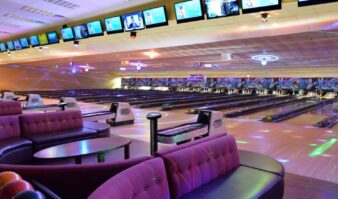 Modern bowling alley with multiple lanes, colorful seating, and overhead screens displaying information. Vibrant lighting creates a lively atmosphere.