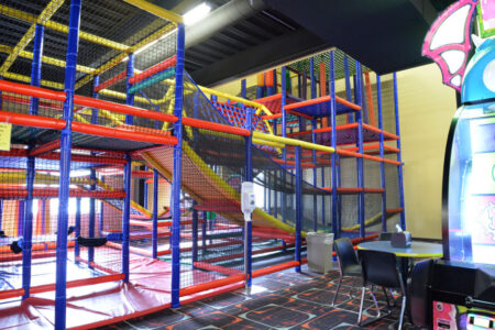 Indoor play area with colorful climbing structures, slides, and nets; nearby are chairs, a table, and an arcade game machine reminiscent of the lively atmosphere at Lanes & Games.