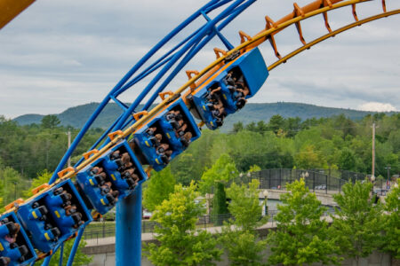 The roller coaster with blue tracks and orange supports twists against a backdrop of trees and hills under a cloudy sky at Six Flags' Great Escape.