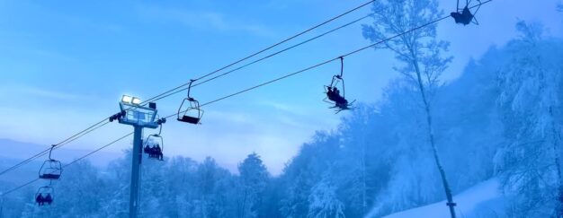 Ski lift with people ascending over a snow-covered landscape at West Mountain, surrounded by frosty trees and a clear blue sky.