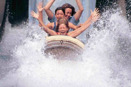 Four people with arms raised are riding a log flume at Hurricane Harbor, splashing through water.